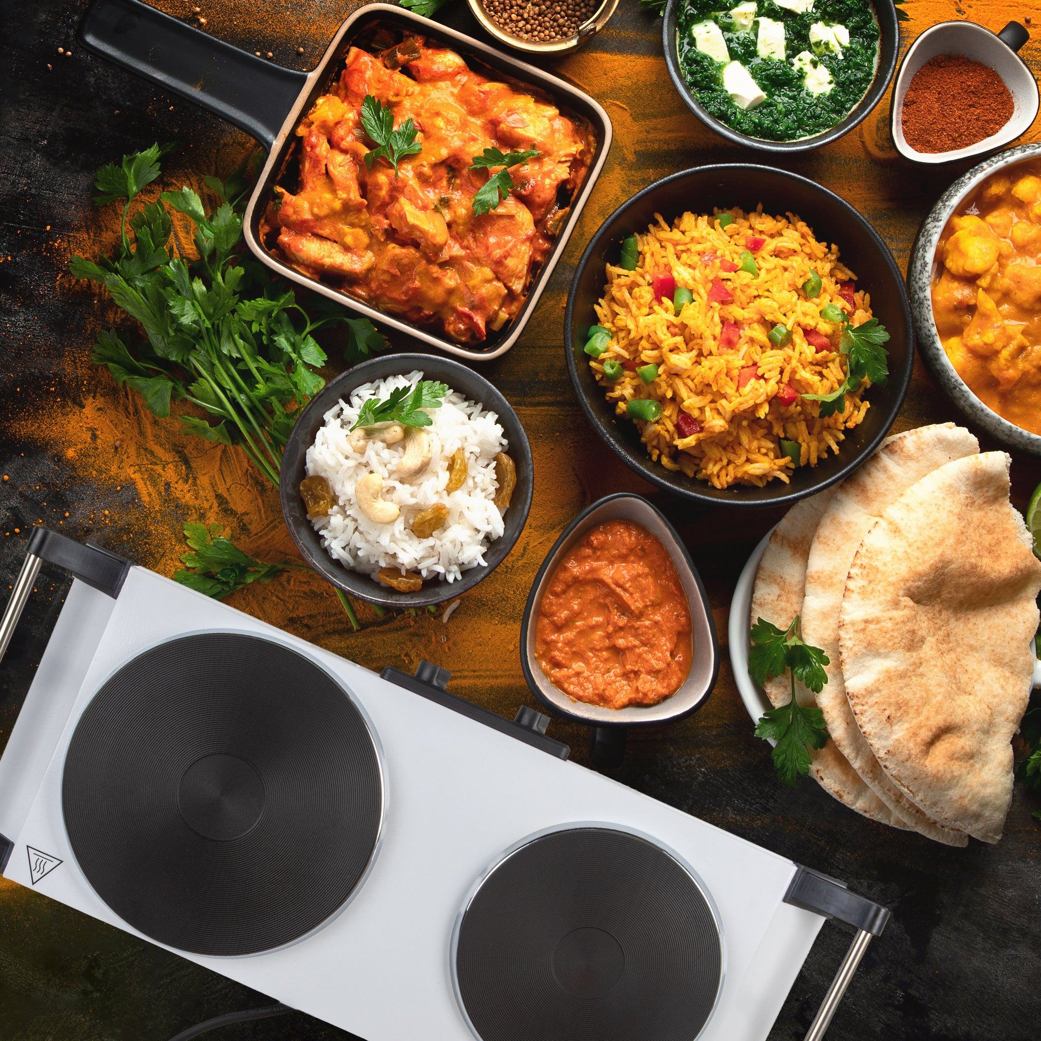 Duronic Electric Hot Plate HP2 WE sitting beside an assortment of Indian curry dishes.