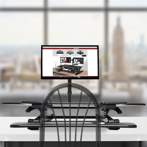 Duronic DM05D5 Corner Sit-Stand Desk with a dual-monitor mount is displayed on a white desk in a bright, modern office overlooking a city skyline.
