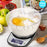 Duronic Digital Kitchen Scale weighing ingredients in a clear bowl with a 5kg maximum capacity, surrounded by baking supplies like eggs, flour, and fresh apples.
