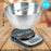 Duronic Digital Kitchen Scale with a stainless steel mixing bowl weighing colorful pasta, including two AAA batteries.
