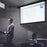 Duronic Electric 60” Projector Screen with a speaker gives a professional presentation in a darkened room using a wall-mounted projector screen displaying digital content.