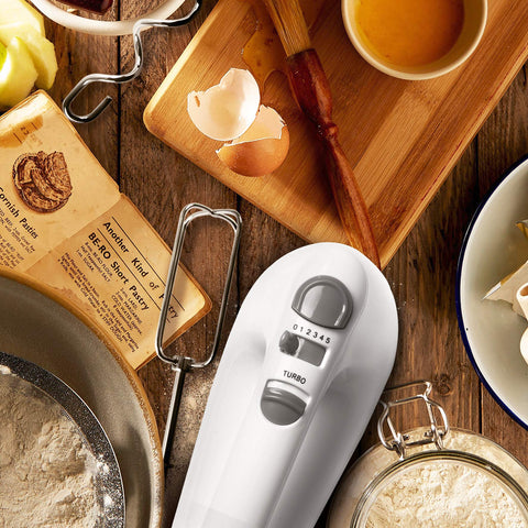 Duronic Hand Mixer Electric Whisk on a wooden table surrounded by baking ingredients, flour, and eggshells.