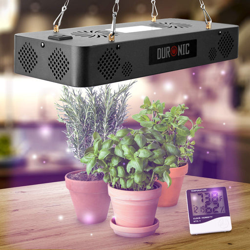 Duronic Hanging Grow Light illuminates a trio of potted herbs including basil and rosemary on a wooden table, accompanied by a digital temperature and humidity monitor.
