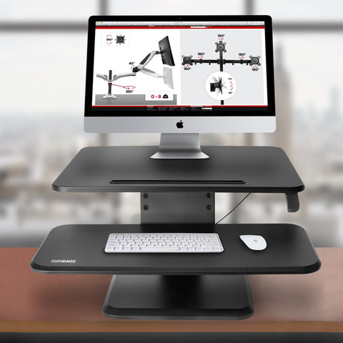 Duronic Sit-Stand Desk featuring a two-tier design with an iMac on the top shelf and a wireless keyboard and mouse on the lower tray.